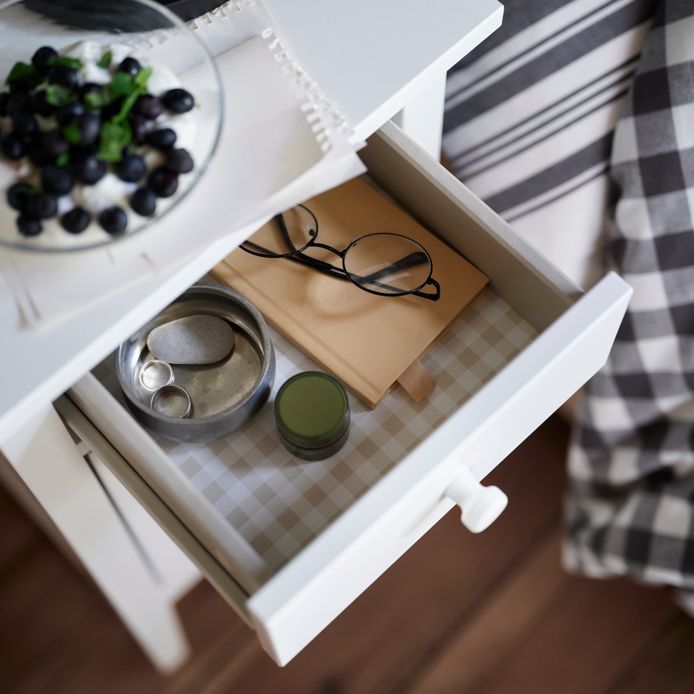 Szafka nocna GULLABERG w kolorze białym z otwartą szufladą, w której znajdują się okulary, notatnik i świeczka, stojąca obok łóżka.