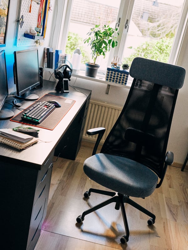 A black swivel chair with armrests and a back in a mesh material stands in front of a desk with a computer on top.