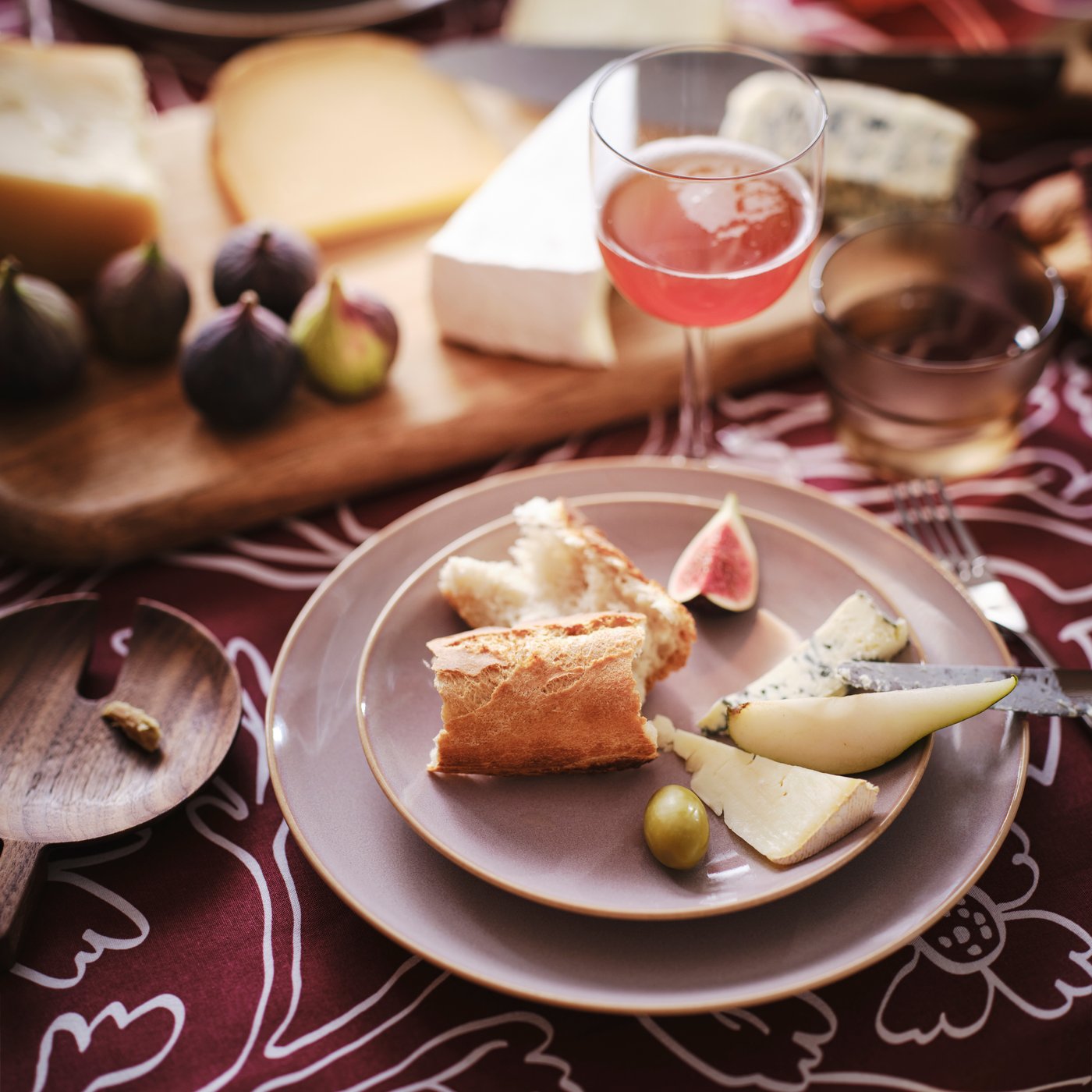 A table set with cheese and figs on a wooden cutting board. FÄRGKLAR plates in light pink hold sliced bread and cheese.