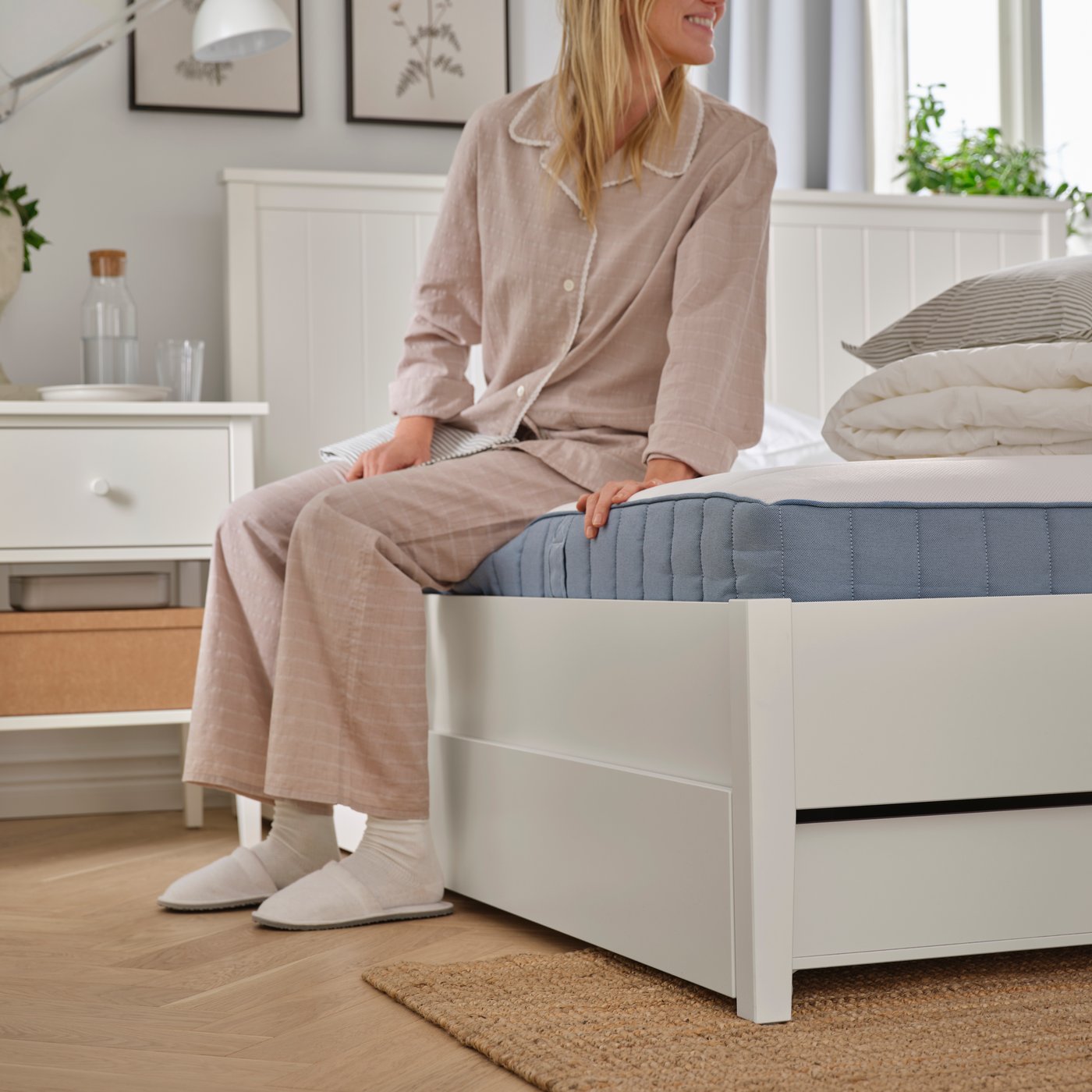 I ett sovrum sitter en person i pyjamas, strumpor och tofflor på en vit GULLABERG sängram med fyra förvaringslådor.
