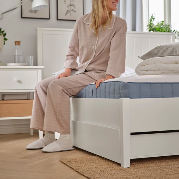 In a bedroom, a person in pyjamas, socks and slippers is sitting on a white GULLABERG bed frame with four storage boxes.