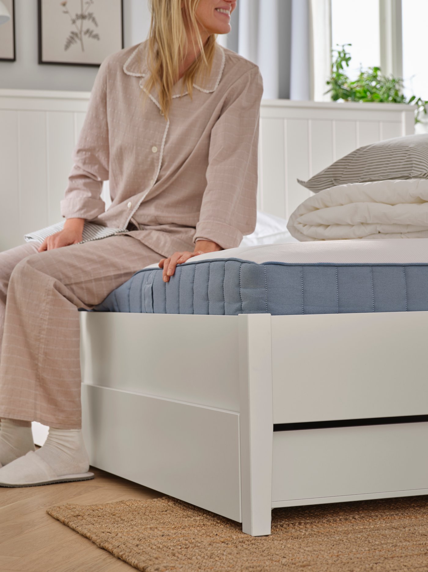 In a bedroom, a person in pyjamas, socks and slippers is sitting on a white GULLABERG bed frame with four storage boxes.