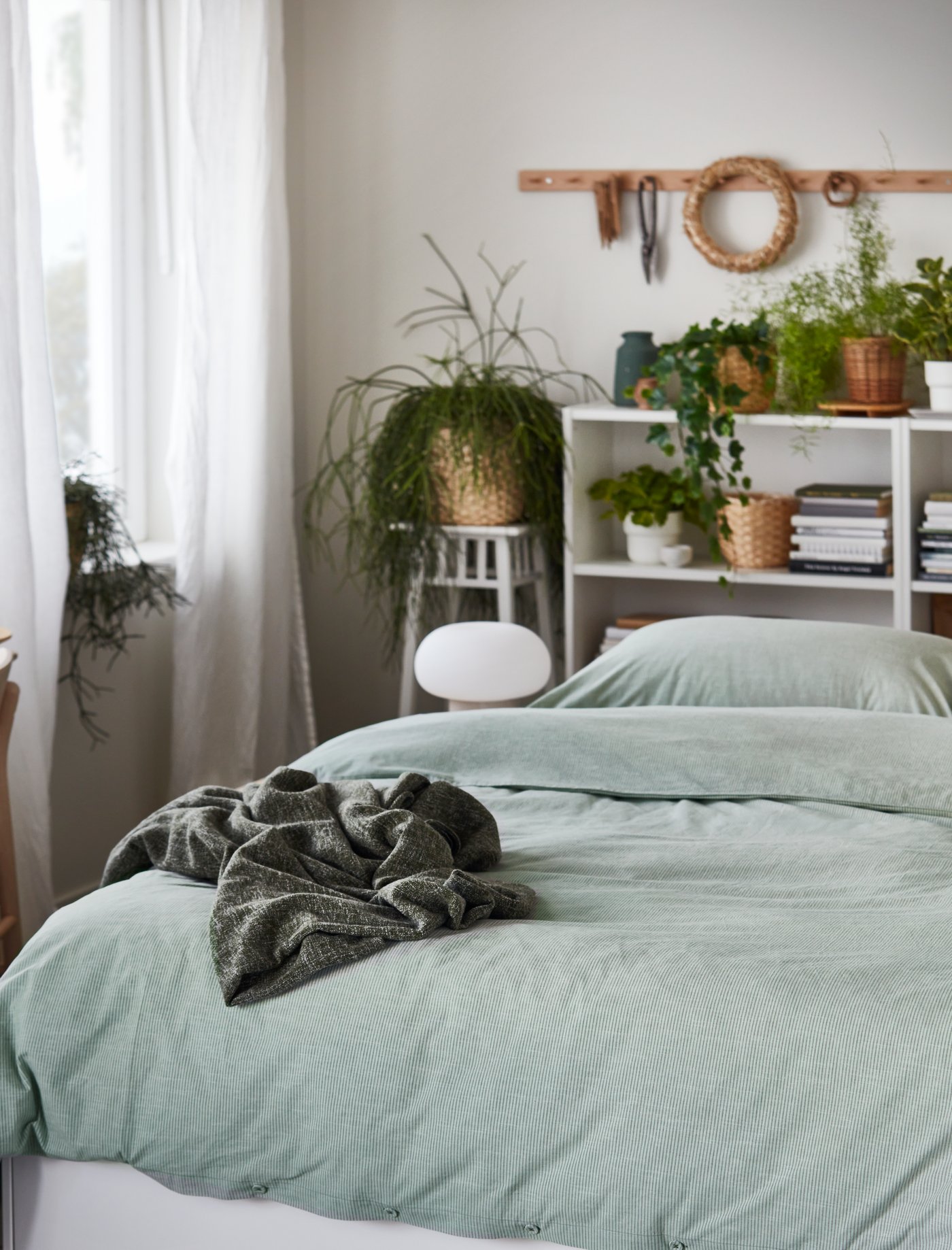 Dormitorio con funda nórdica verde y 2 fundas de almohada de algodón con una manta gris encima y una estantería blanca.