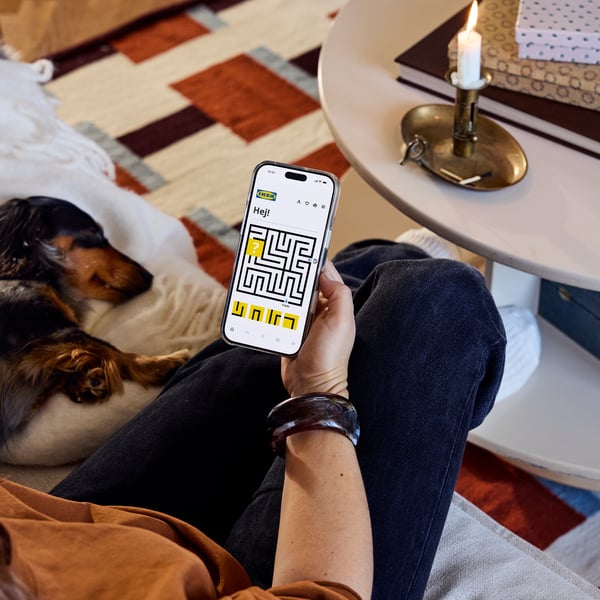 Osoba siedzi na kanapie z psem, trzyma telefon z aplikacją IKEA i żółtym labiryntem.