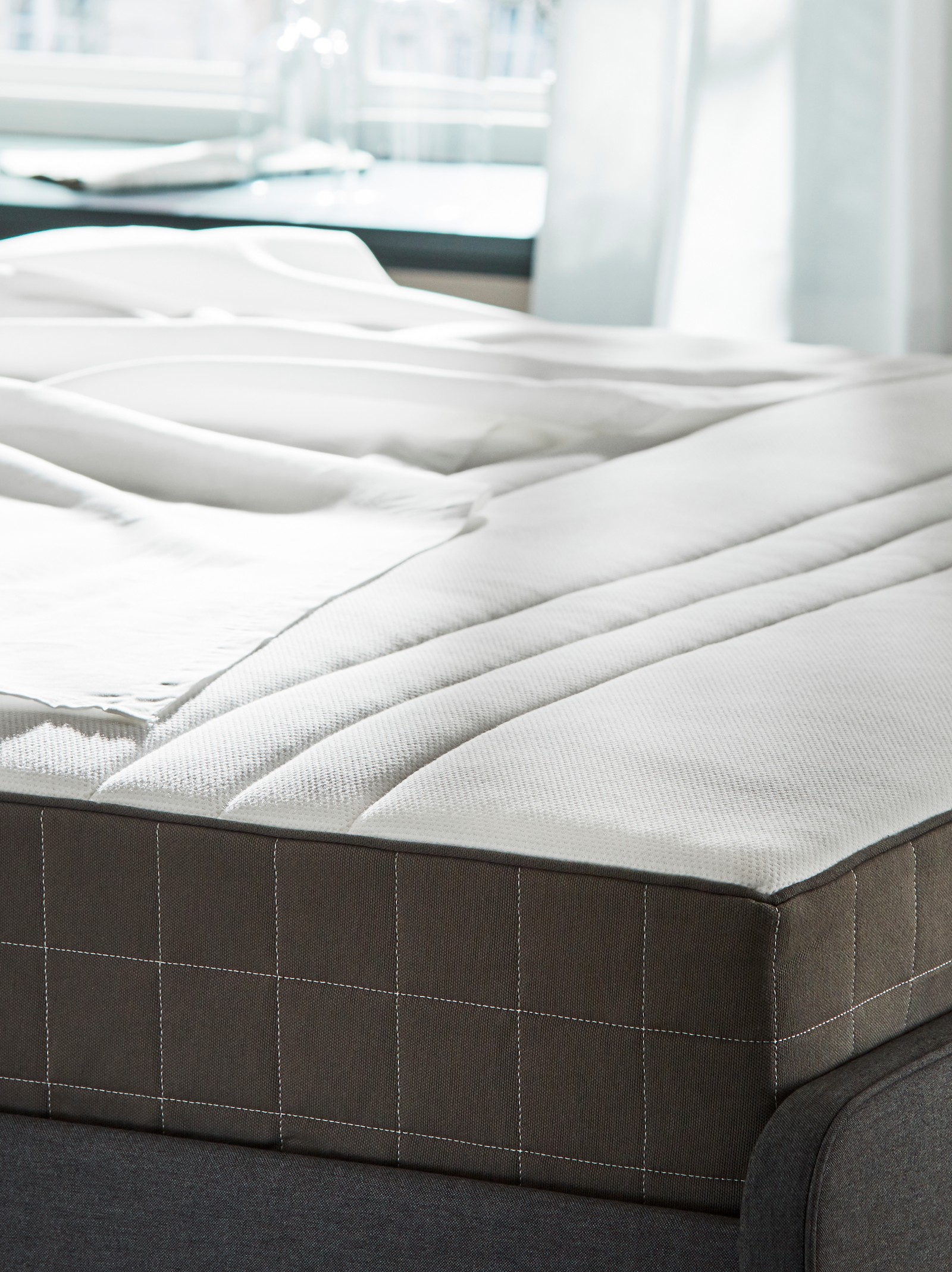 На кровати с обивкой ХАУГА лежит матрас с пружинами карманного типа ХОВОГ. Кровать стоит в спальне перед окном.