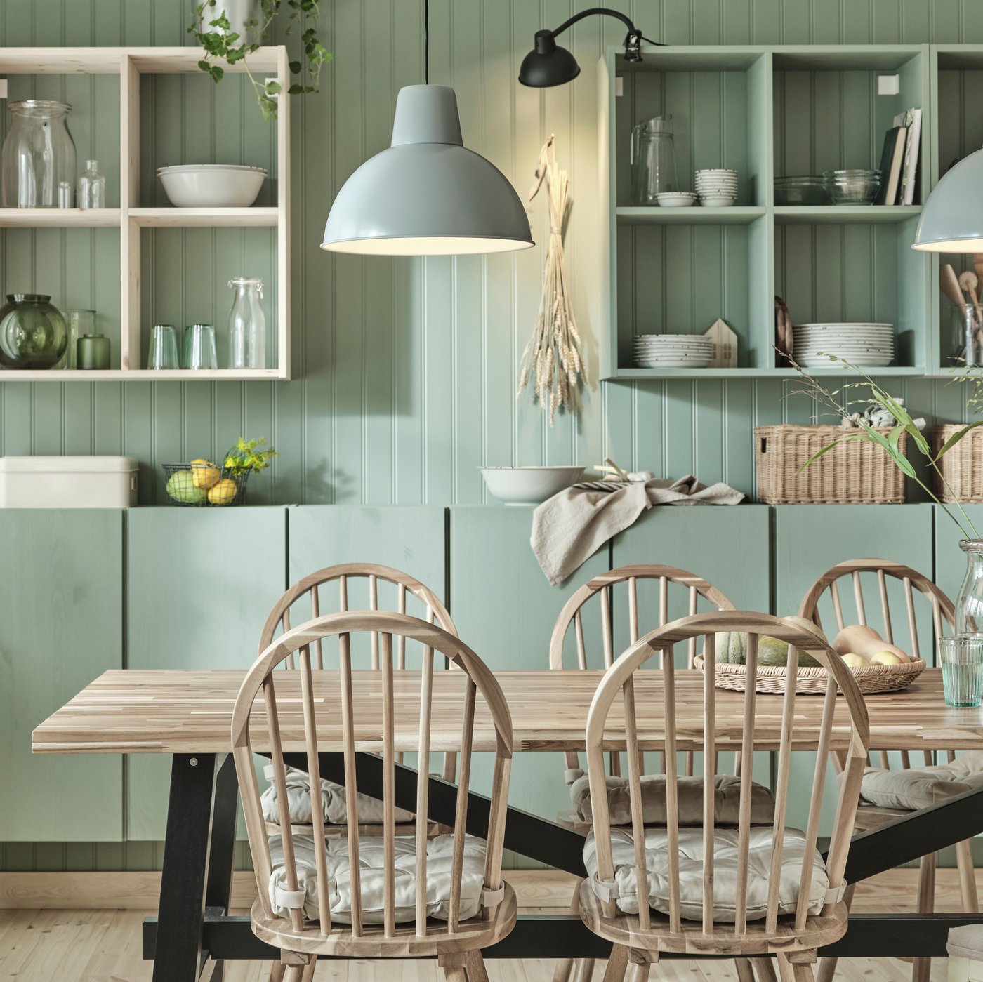 A wooden SKOGSTA dining table and matching SKOGSTA chairs stand in a dining room in front of green-painted IVAR cabinets.