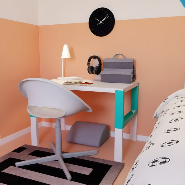 Espacio de estudio con escritorio blanco PÅHL , silla moderna, lámpara, libros y organizador, junto a una cama con ropa de cama de balones de fútbol y una pared naranja con reloj negro