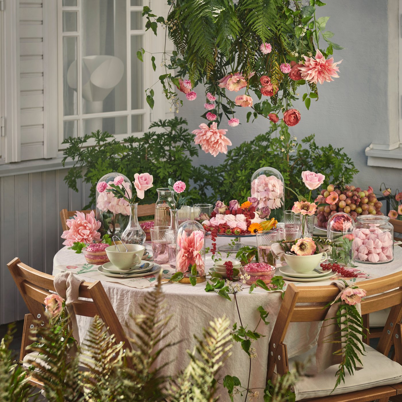 Runt bord utomhus dukat med rosa blommor, glasburkar med godis och porslin, omgiven av trästolar och gröna växter med hängande blommor ovanför.