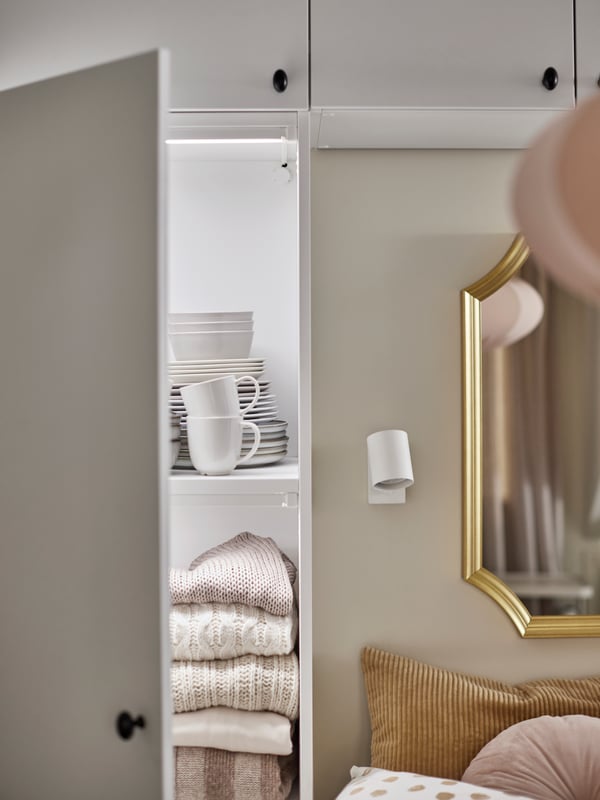 A white PLATSA wardrobe system beside and above a bed, with one door open showing tableware and folded sweaters inside.