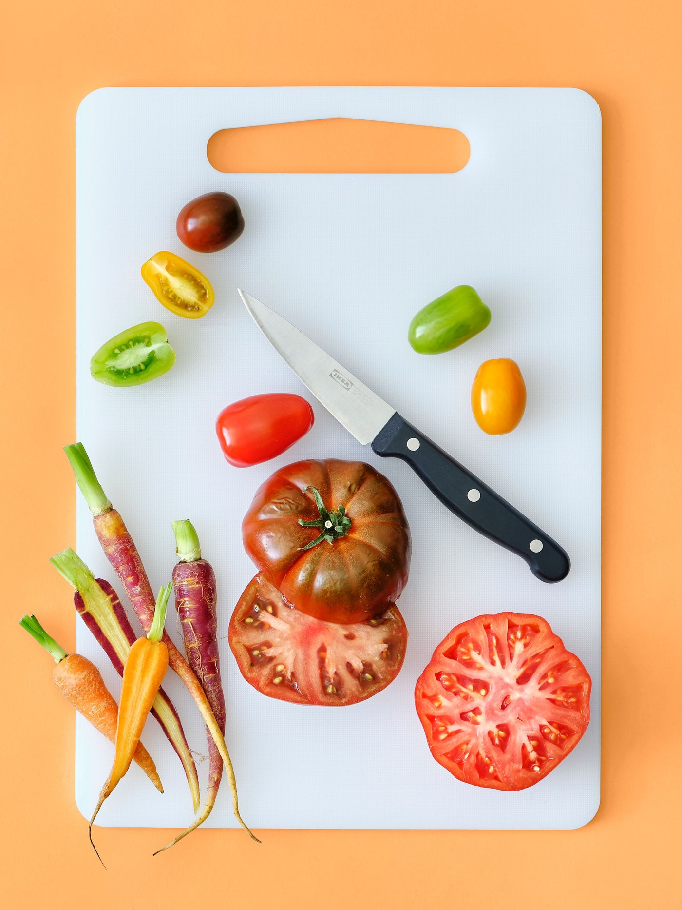 A VARDAGEN cook’s knife is placed on top of a white LEGITIM chopping board with carrots and tomatoes in different colours.