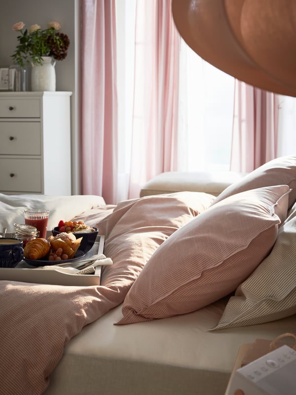 A tray with a breakfast spread on a bed with a pink duvet cover and pillows in pink and grey pillow cases.