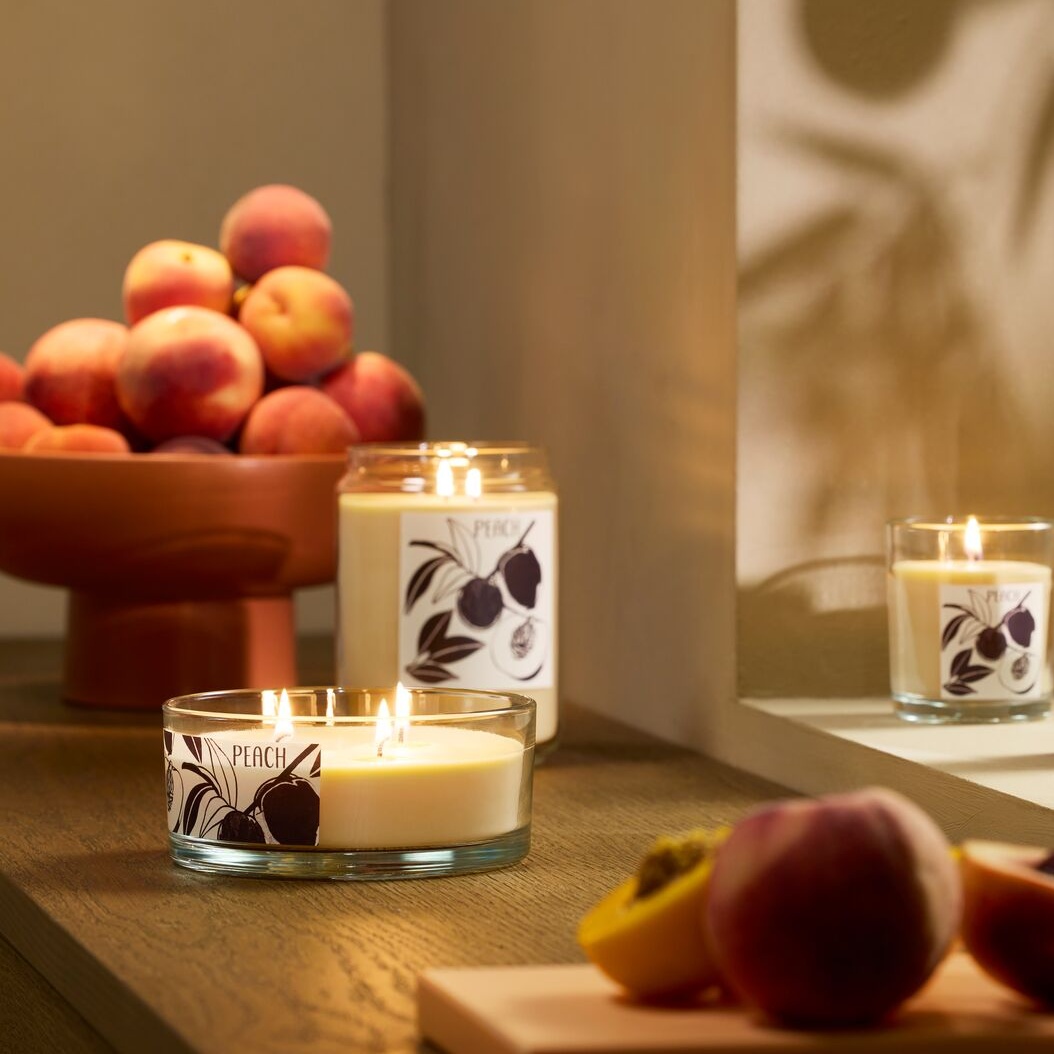 Three lit peach-scented candles with peaches in the foreground and background.