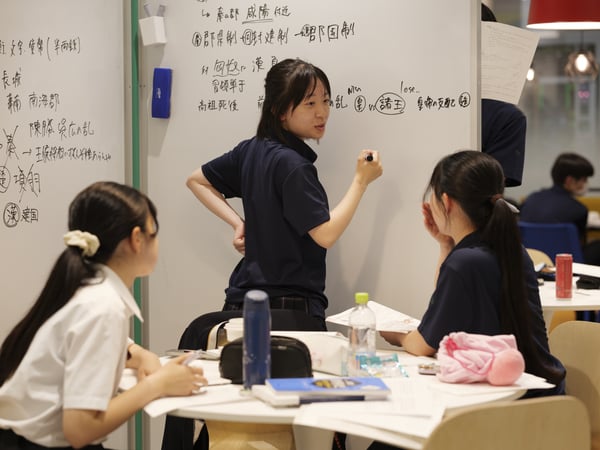 ホワイトボードを使って議論する3人のチーム。机上にはノートや文房具が並ぶコラボレーションスペース