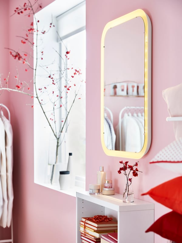 Un espejo con luz STORJORM de bordes redondeados en un baño con muros rosado claro, instalado sobre un estante blanco y estrecho junto a una ventana. 