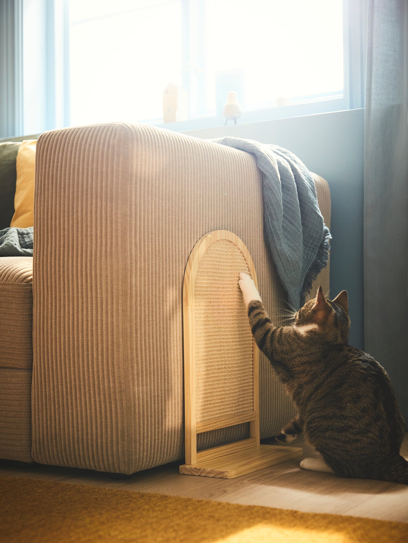 Katt klöser på ett UTSÅDD klösträd som är monterat på sidan av en beige soffa med filt över armstödet i ett solbelyst rum.
