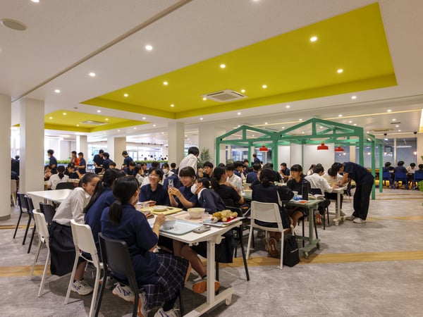 明るい黄色の天井パネルの下で、多くの人がテーブルに座って交流している広々とした室内