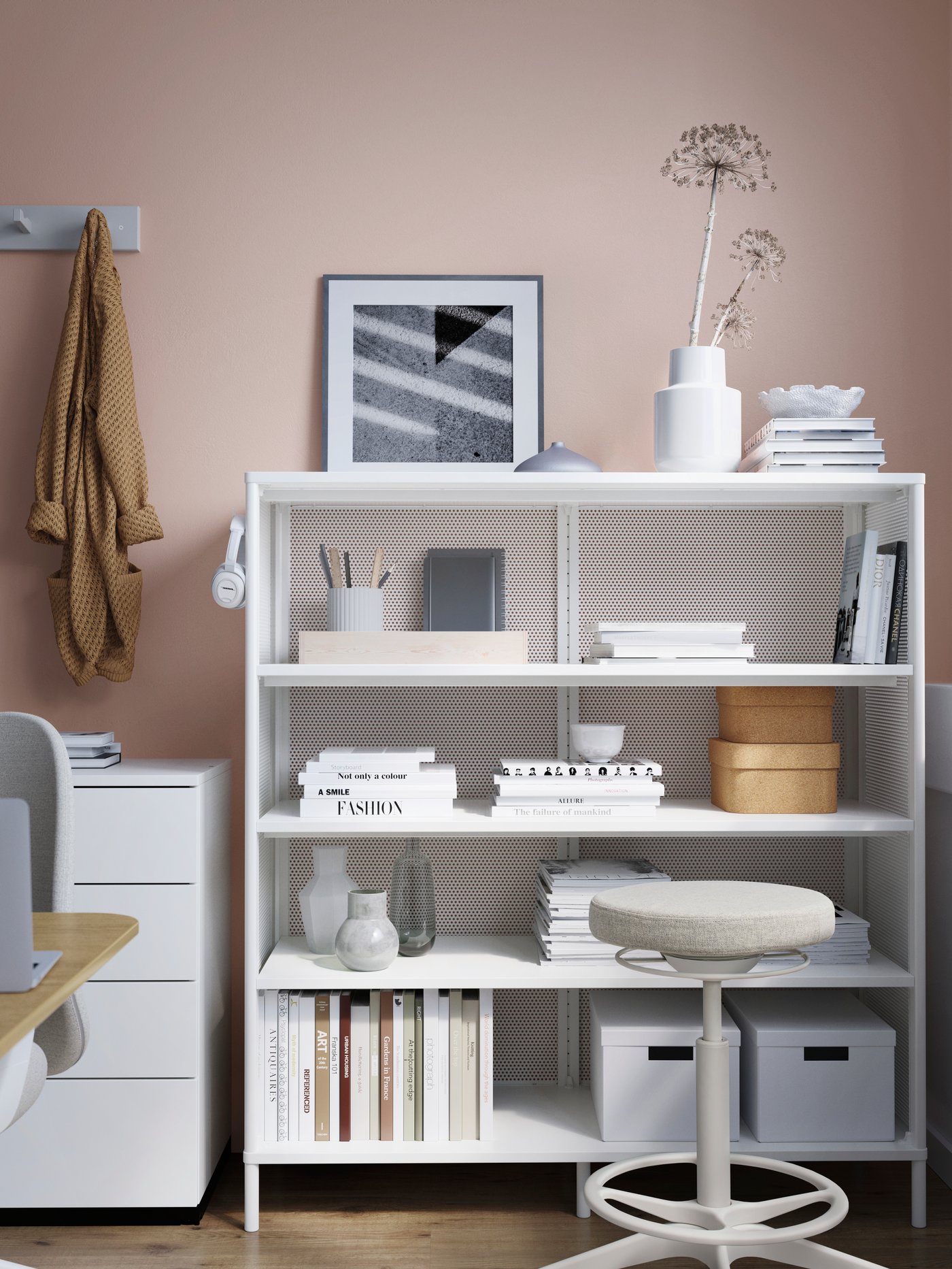 A white BEKANT shelving unit holding books, vases, stationery, headphones and storage boxes. A cream stool is in front.