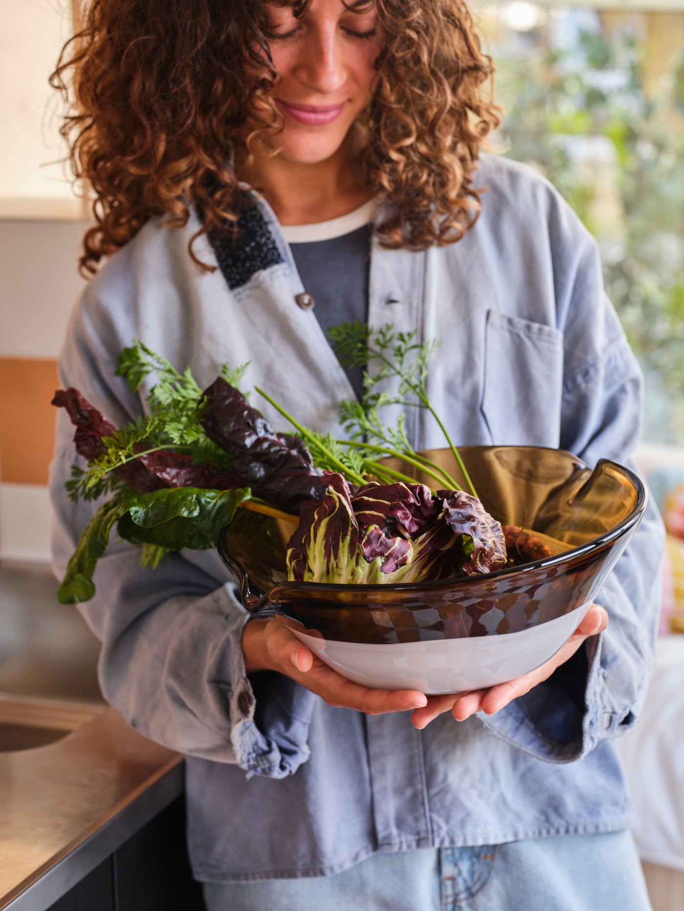 A person holds an OMMJÄNGE double-spouted serving bowl in brown/white glass in front of them filled with various vegetables.