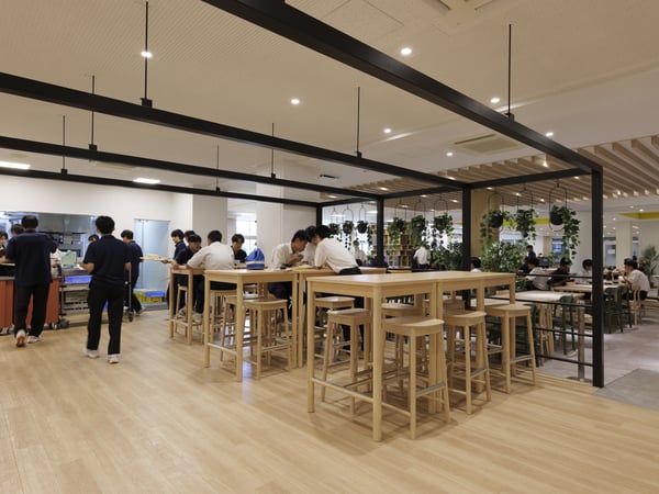 高テーブルとスツールが並ぶ交流スペースで、数人が会話や食事を楽しんでいる様子