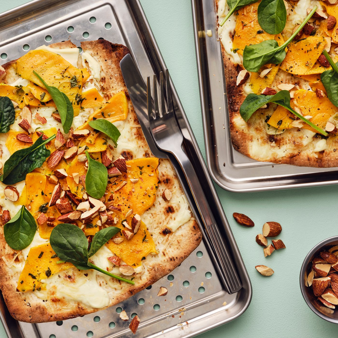 Two flatbread pumpkin pizzas topped with spinach and MUNSBIT roasted almonds on stainless steel trays with cutlery.