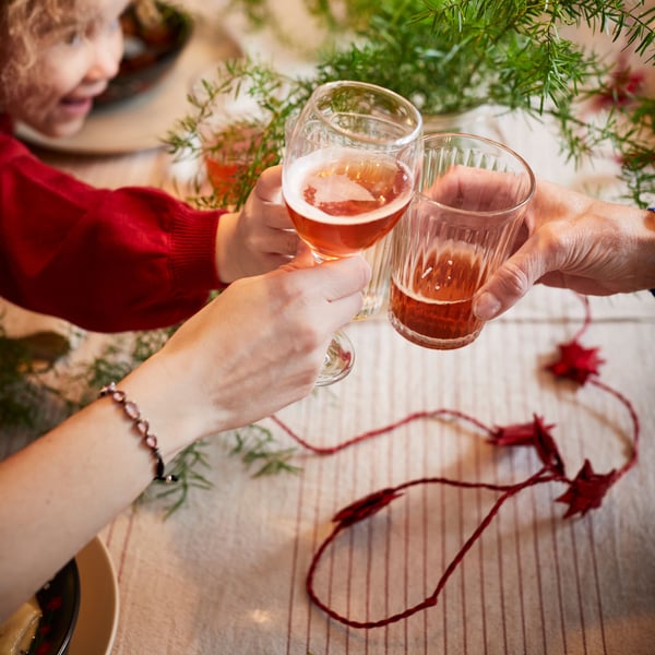 Manos sosteniendo vasos VARDAGEN y ÄNKEBLOMSTER con bebestibles de fiestas y decoración navideña.