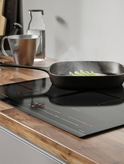 Close-up of a black induction hob, on which is a black grill pan with steaming food. Next to the pan is a carafe with lid.