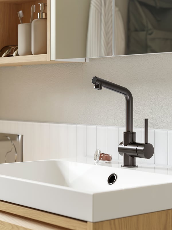 A close-up of a white ORRSJÖN wash-basin with a black LUNDSKÄR wash-basin mixer tap and a watch placed next to it.