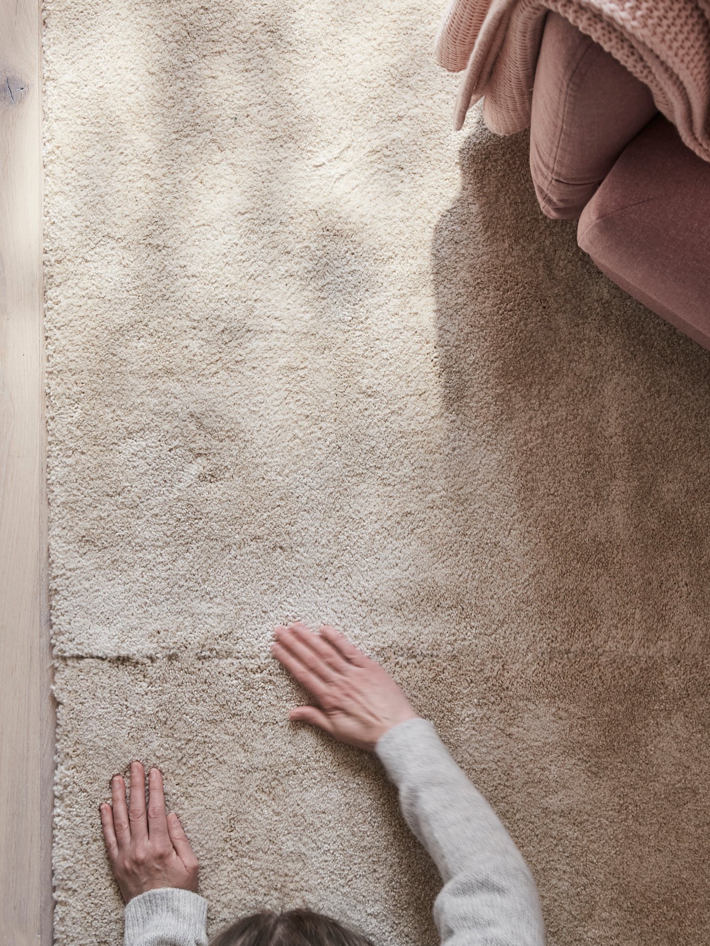 Una vista aérea de una persona ajustando dos alfombras de pelo bajo rectangulares STOENSE en color hueso junto a un sillón parcialmente visible.