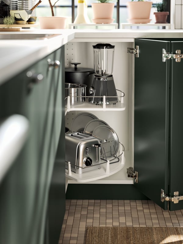 A cabinet with deep green kitchen fronts and doors open, showing an UTRUSTA corner base cabinet carousel with various items.