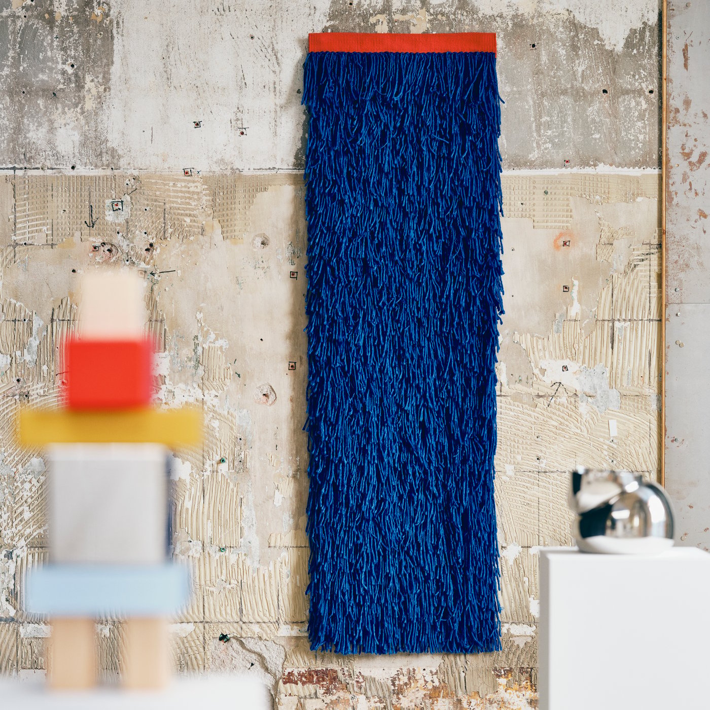 Close up of three objects from IKEA KONSTRUNDA art collection. A blue rug with long lashes is mounted against a concret wall.