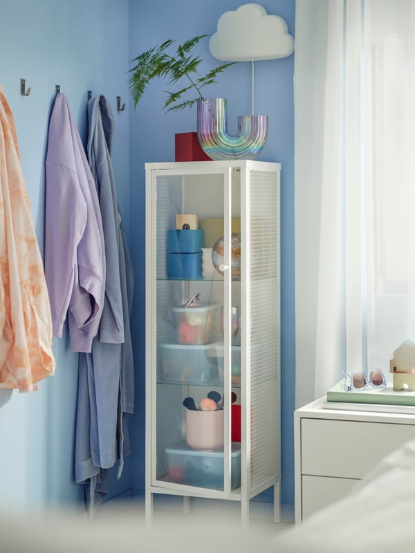 In einem blauen Raum steht eine weiße Vitrine an der Wand mit Deko darin. An der Seite hängen bunte Handtücher.