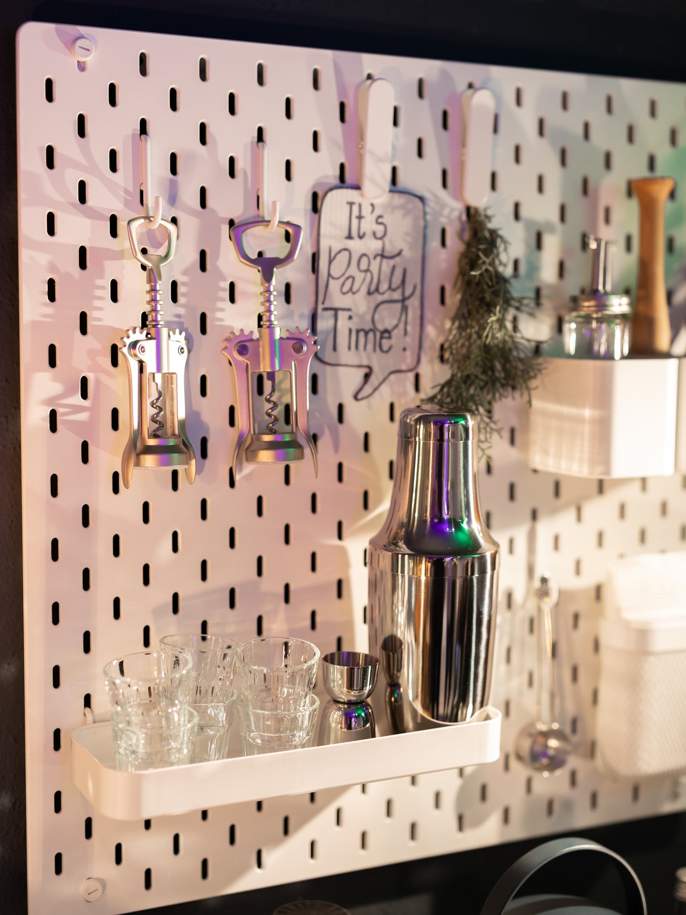 A white SKÅDIS pegboard displaying various bar essentials such as a corkscrew, a cocktail shaker, and glassware.