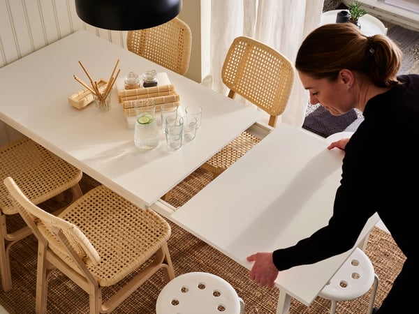 Mujer extendiendo una mesa extensible VANGSTA blanca sobre una alfombra de yute, con sillas de bambú claras y pisos para sentarse blancos a ambos lados.
