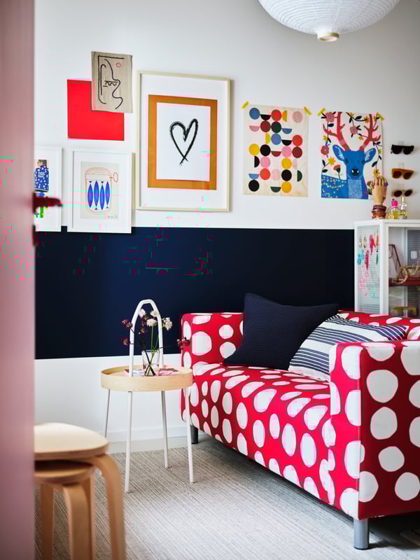 By a blue and white wall filled with art stand a red-and-white, polka-dot KLIPPAN two-seat sofa and a BURVIK side table.