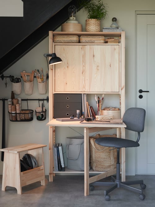 Eine IVAR Aufbewahrung mit Schreibtisch unter einem Treppenabsatz. Ein Drehstuhl ist seitlich zu sehen. Stangen mit Behältern an der Wand sind der Aufbewahrungsort für Werkzeug. Auf dem Schreibtisch steht eine Arbeitsleuchte.