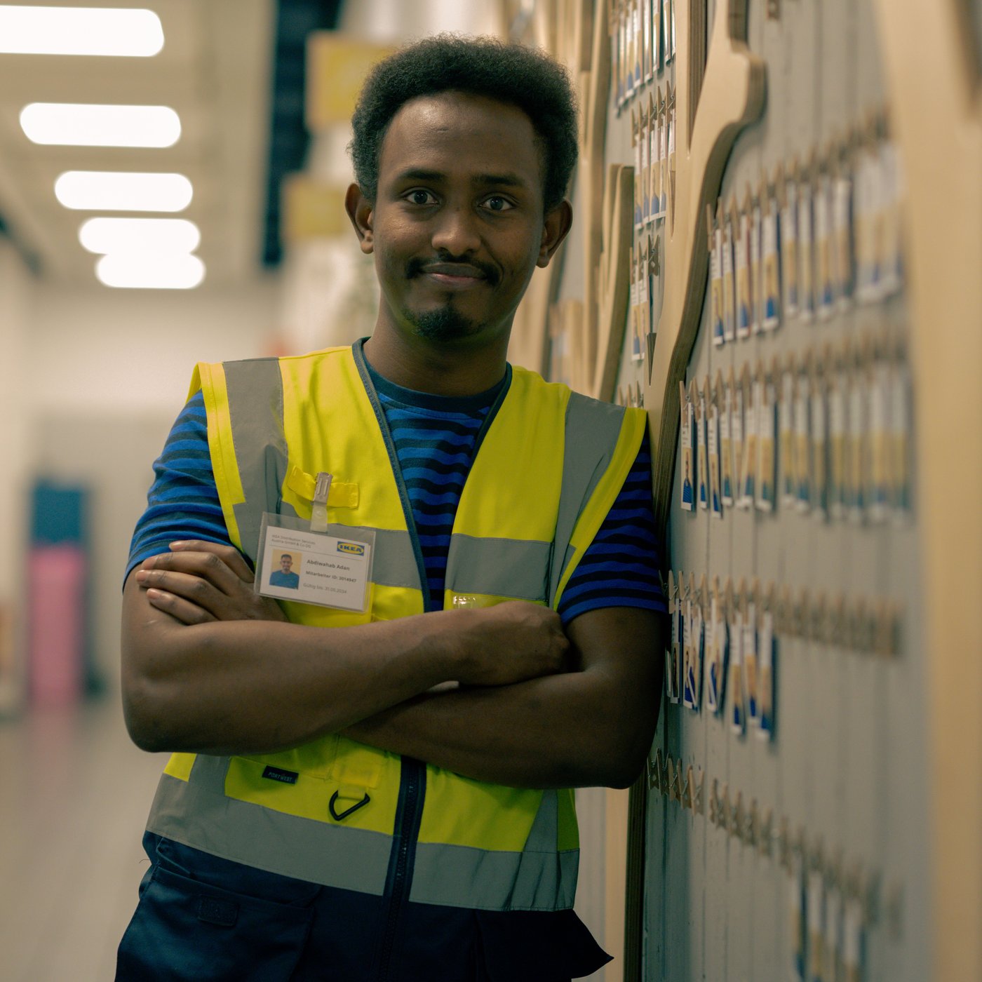 Abdi, wearing a high-visibility vest, stands with arms crossed in a corridor, smiling.
