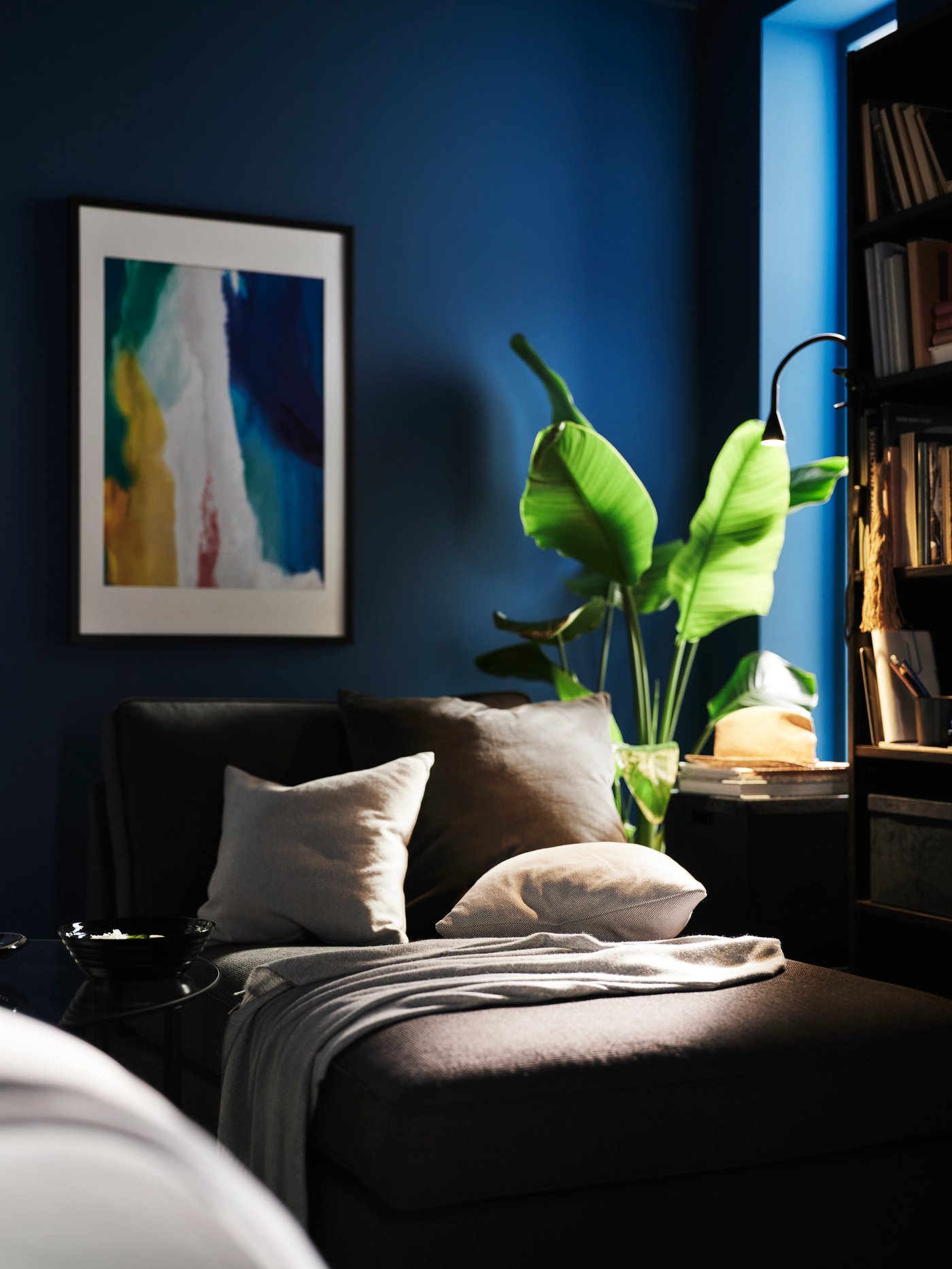 En antracitgrå KIVIK schäslong i ett blått rum med olika kuddar och en pläd ovanpå. Bakom står en Vogar BILD affisch.