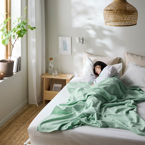 A woman sleeping in bed in an all white/beige bedroom with the light shining through a window at the left.