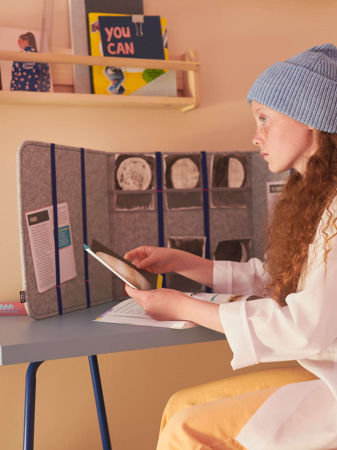 A teenage girl in a beanie sitting at a blue LAGKAPTEN/TILLSLAG desk with an ÖVNING desk divider placed on it.