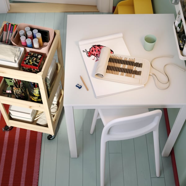 In uno spazio di lavoro ordinato e luminoso, un tavolo VIHALS bianco attorno al quale ci sono una sedia e accessori per le attività artistiche organizzati su un carrello.