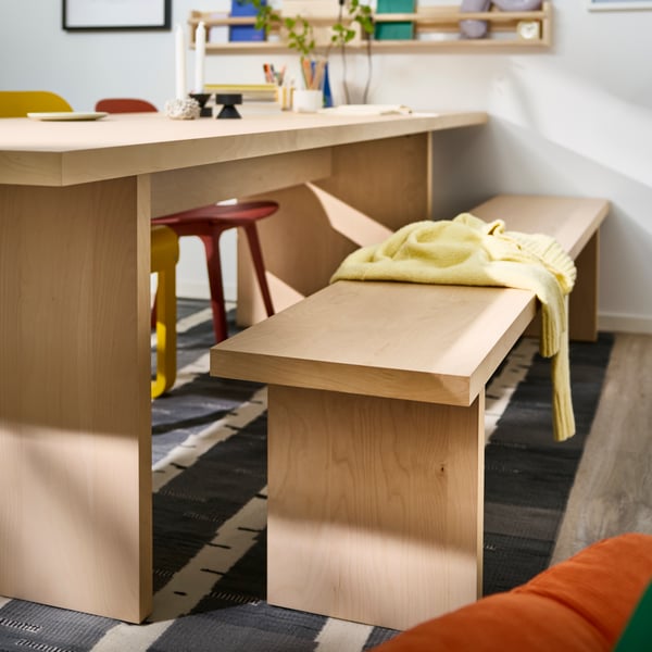 A BERGSHYTTAN bench in bright birch veneer is standing next to a large BERGSHYTTAN table in the same style and colour.