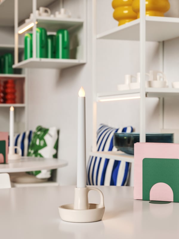 Wall-mounted white PLATSA open shelving units display colourful home deco objects. There are LED candles on the white tables.