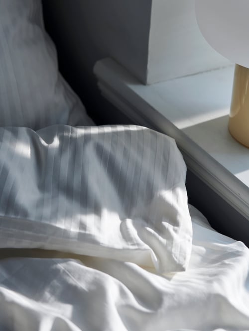 Ein ungemachtes Bett mit der weißen NATTJASMIN Bettwäsche, daneben ist eine eine DEJSA Tischleuchte zu sehen.