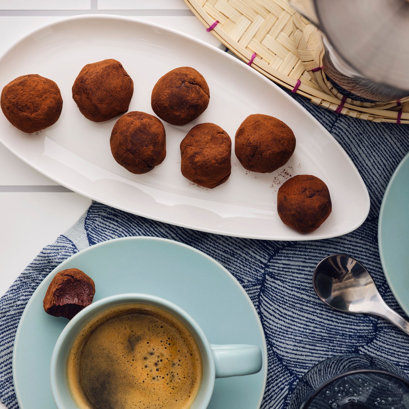 A white leaf-shaped plate with seven chocolate truffles, placed next to two turquoise cups on saucers with coffee.