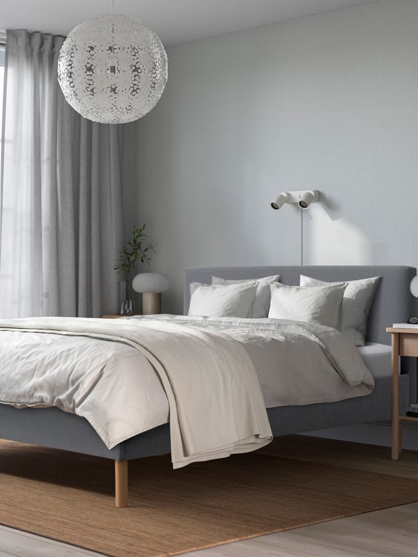Ein gepolstertes NARRÖN Doppelbett in grau mit Holzbeinen. Darauf liegt Bettzeug und eine Tagesdecke. 