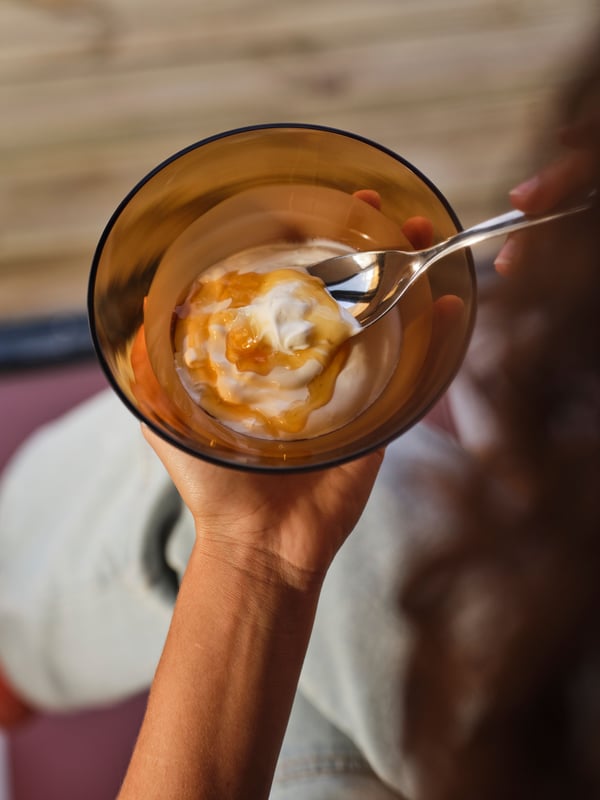 An OMMJÄNGE bowl made of brown/white glass rests in one hand, while another hand holds a spoon scooping up food from the bowl.
