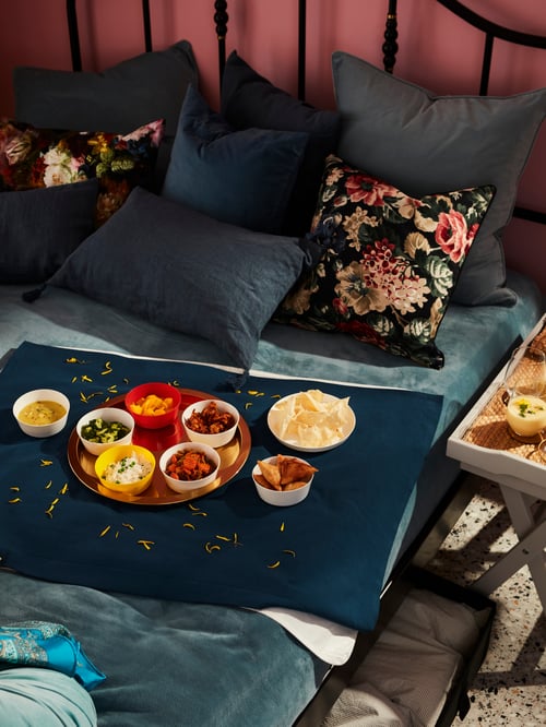 A made bed has a thin cushion on it like a table. A round, brass-coloured GLATTIS tray with bowls of food is on the cushion.