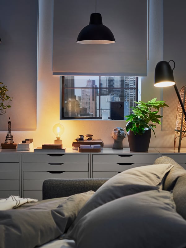 VIMLE sofa-bed as a bed, lit by a SKURUP floor/reading lamp, with four, white ALEX drawer units in the background.