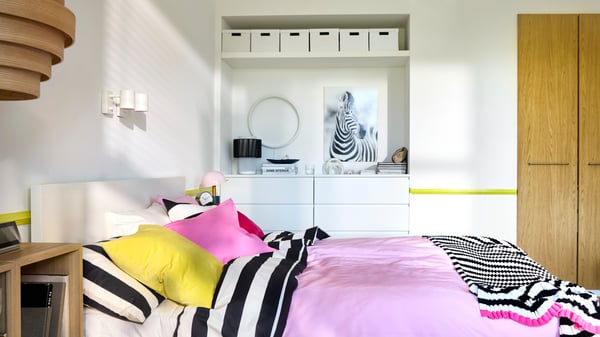 A bedroom with white walls, built-in storage in the back and a double bed with bright pink and black/white striped bedding.