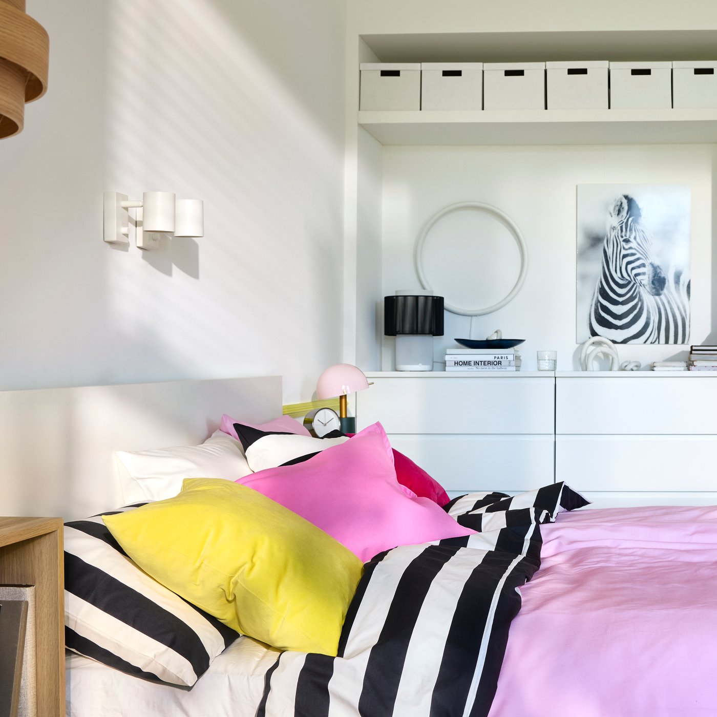 A bedroom with white walls, built-in storage in the back and a double bed with bright pink and black/white striped bedding.